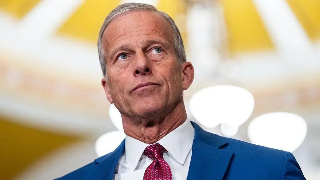 John Thune is pictured from a low angle, wearing a blue suit and red tie. The background of an indoor hall is blurred.