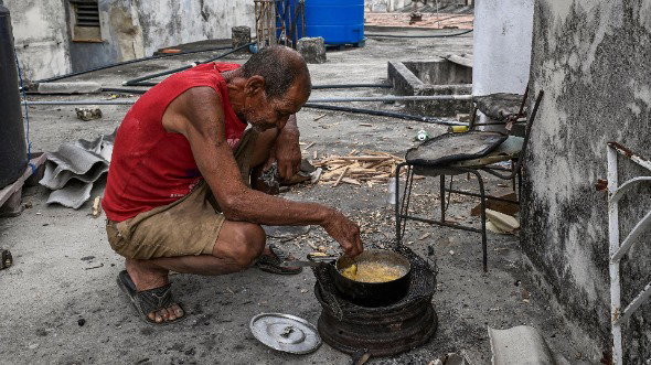 A man crouches to cook with firewood on the ground in Havana. There is debris on the ground around him.