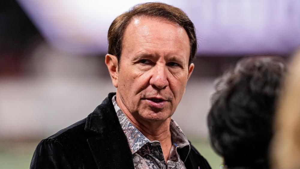 Jeff Landry, wearing a dark jacket and floral shirt. The background is blurred and he has short brown hair.