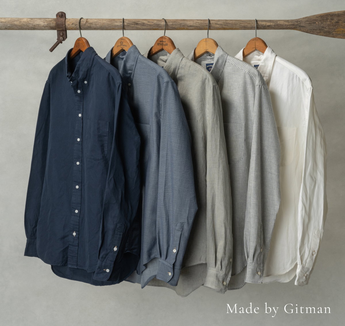 Five Bills Khakis Standard fit shirts hang from hangars on a wooden oar against a light grey colored backdrop. Small text overlay reads 'Made by Gitman'