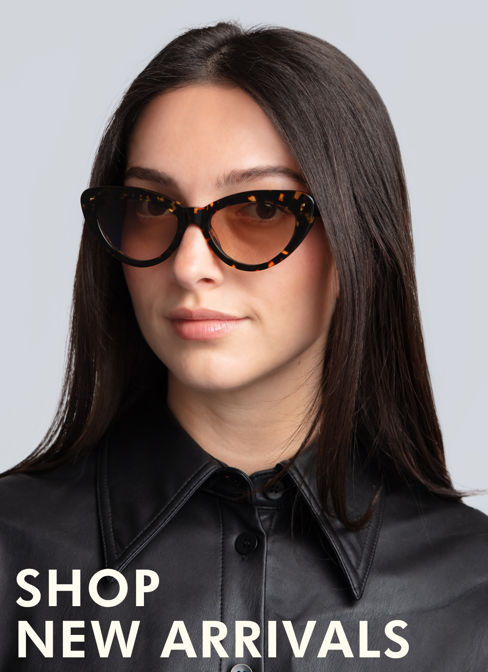 A headshot of a woman with long, straight dark brown hair wearing bold cat-eye sunglasses in a dark tortoise-shell pattern with warm brown gradient lenses. She is wearing a black leather button-up shirt. Text in the lower-left corner reads: 