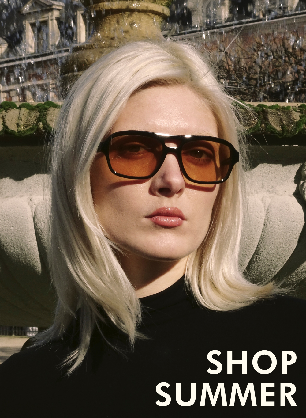 A campaign shot of a woman with platinum blonde hair standing outdoors in front of a stone fountain. She is wearing oversized aviator-style sunglasses with dark frames and bright orange lenses. Text in the lower-right corner reads: 