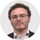 A headshot of Jack Fenwick, who has short dark hair and facial hair. He wears glasses, a white shirt and blazer.