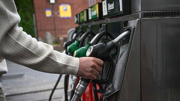 A person, wearing a white jumper, places their hand on a petrol pump in a forecourt in a nondescript location in the UK.