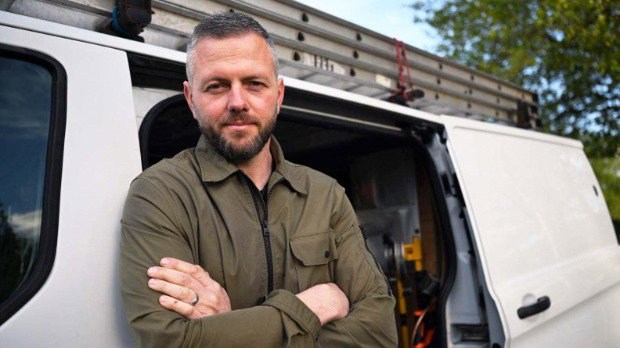 Robert Kenyon stands with his arms crossed, leaning aginst a white van. He is wearing a khaki coloured jacket.