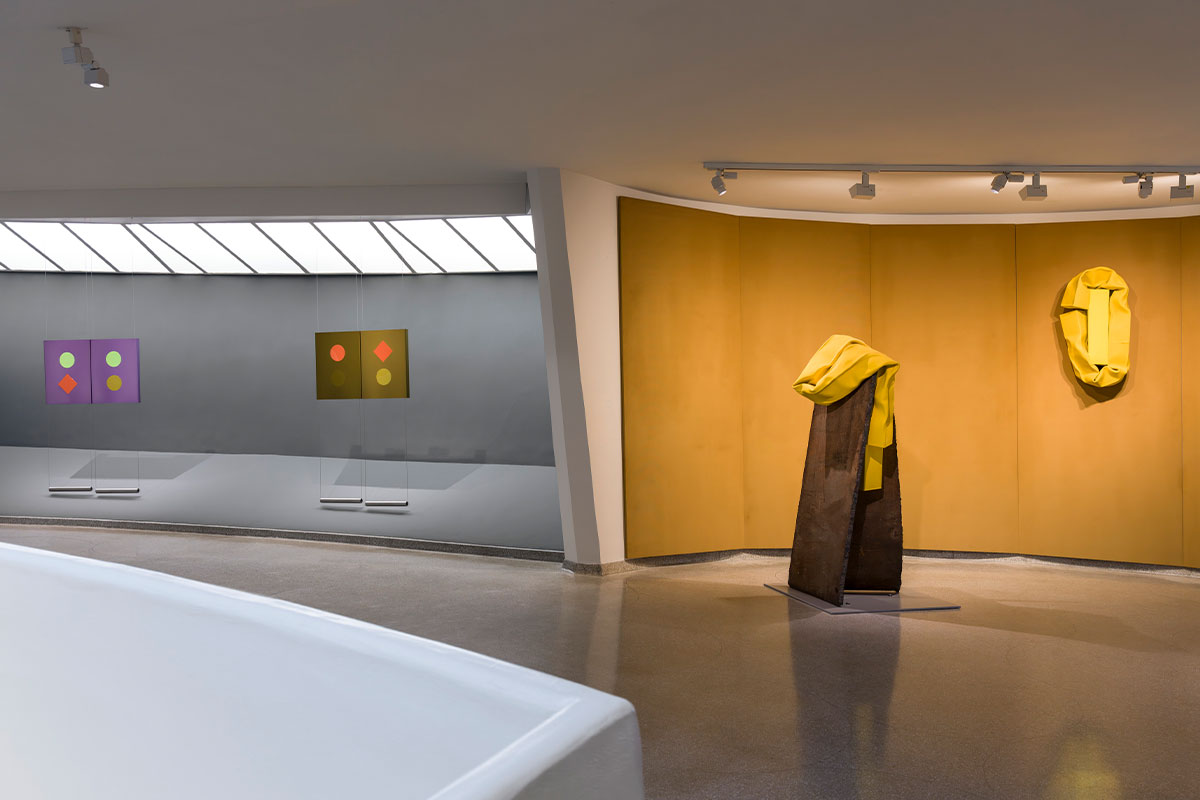 Interior of the Guggenheim Museum with abstract geometric paintings on the left and two yellow‑draped sculptures on a mustard wall to the right, under soft track lighting.