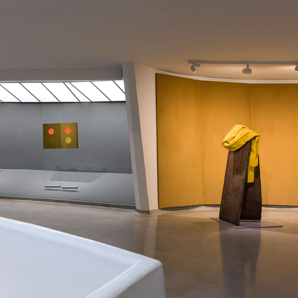 Interior of the Guggenheim Museum with abstract geometric paintings on the left and two yellow‑draped sculptures on a mustard wall to the right, under soft track lighting.