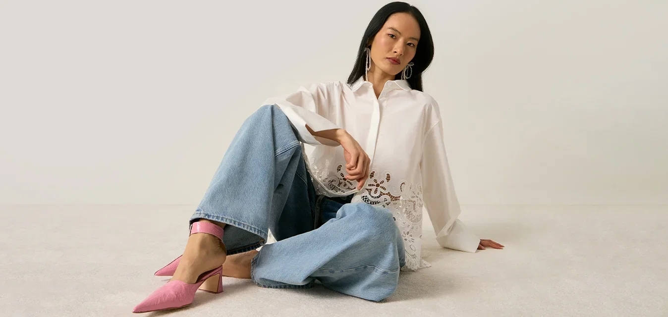 Vrouw zittend op de vloer, gekleed in een wit kanten hemdje, jeans met wijde pijpen en roze schoenen met hakken, tegen een effen lichte achtergrond.