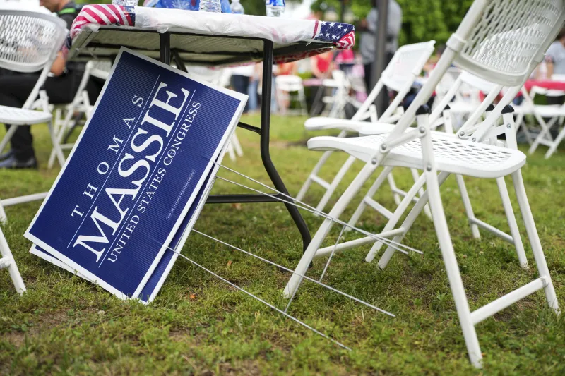 Campaign signs for Thomas Massie sit underneath a white outdoor table and chairs on grass.