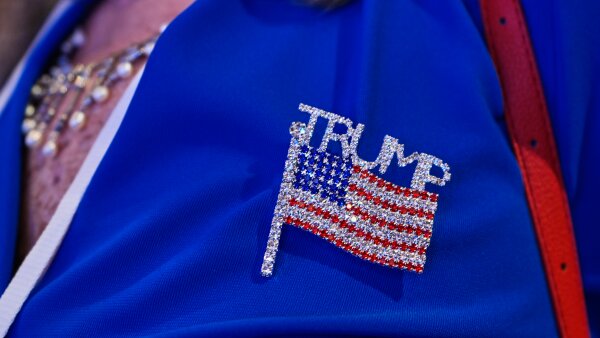 A close-up look at a person’s blue jacket that has a bedazzled Trump pin that reads Trump above an American flag. 