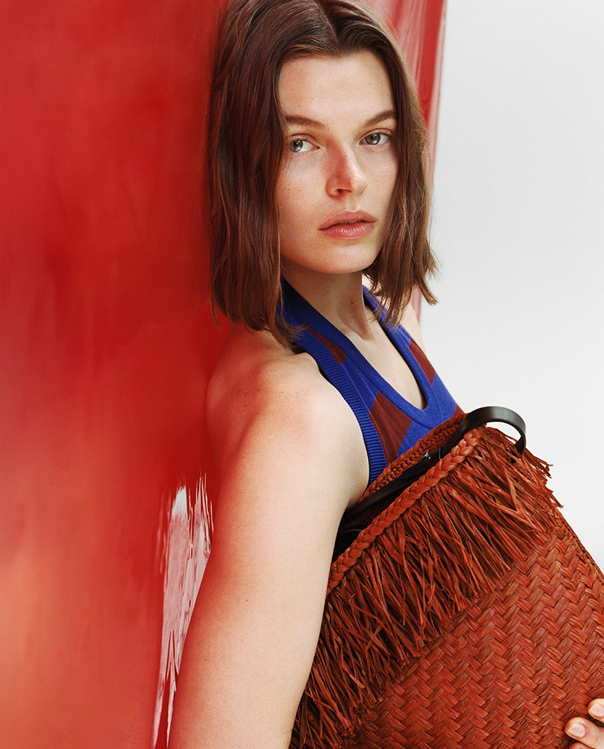 Cropped image of model standing against a red vinyl background, wearing the Frann Dress in Cotton Stripe in Sapphire Multi with the Bucket Bag in Raffia in Rust