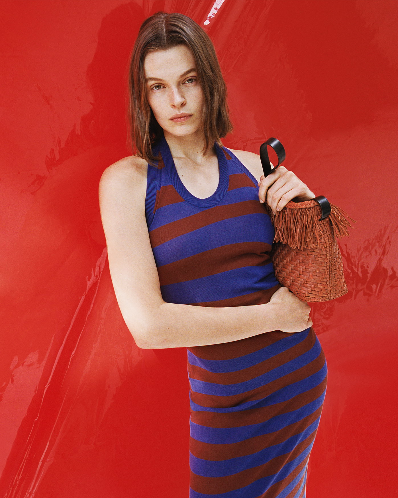 Cropped image of model standing against a red vinyl background, wearing the Frann Dress in Cotton Stripe in Sapphire Multi with the Bucket Bag in Raffia in Rust