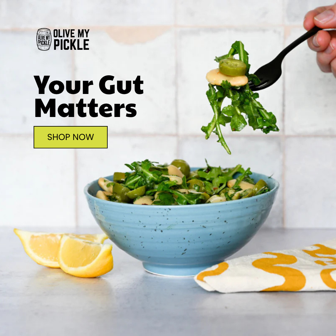 A hand using a fork to lift a bite of fresh salad with arugula, white beans, and green olives from a blue ceramic bowl. Sliced lemons and a yellow-patterned nap