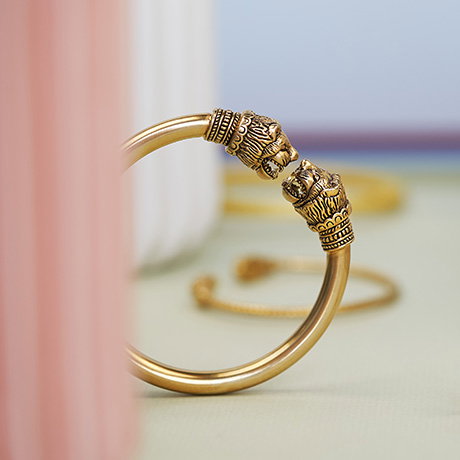 Gold-plated bracelet featuring two lion head finials emerging between ceramic pillars against a soft pastel background. - Shop Jewellery.