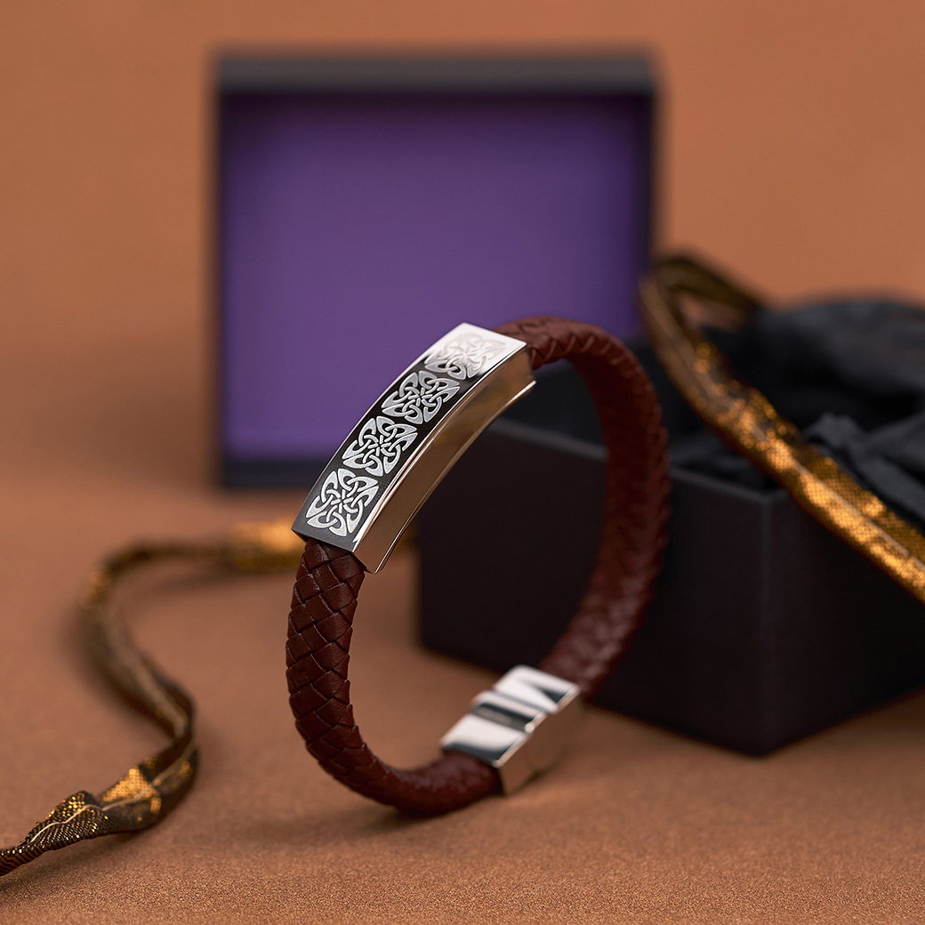 Handcrafted brown leather men’s bracelet featuring Celtic knotwork designs set on a copper-brown background with a gift behind it. - Shop Inspired by Celtic Art. 