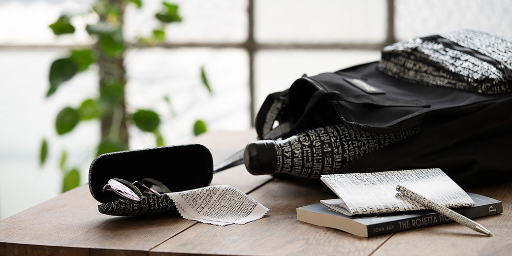Selection of Rosetta Stone-inspired gifts, including a backpack and water bottle featuring white script on a black background, displayed on a table. - Shop ‘Inspired by…’