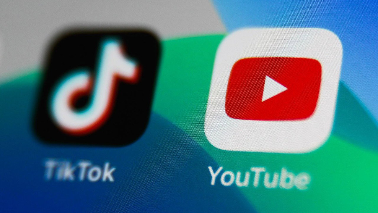 The logo for two apps are close-up on a screen. TikTok is shown with a white and navy musical note icon and YouTube is shown with a red play button icon. The screen background is blue and green.