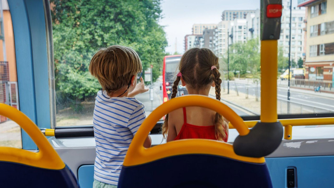 Two children face away from the camera looking out of the top story of a double-decker bus. The bus has yellow grab-handles.