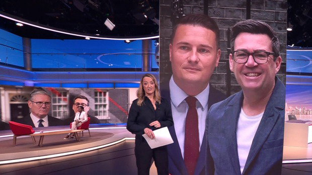 Laura Kuenssberg stands, dressed all in black with long, blonde hair, in a studio. To her left is a large picture of Andy Burnham and Wes Streeting and her three person panel, and to her right is an interviewee, Kemi Badenoch, sat by a wooden table.