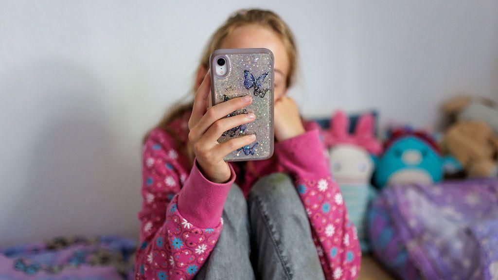 An anonymous young girl looking at her phone on her bed in a room with brightly coloured cuddly toys. She is wearing a pink flowery jumper and jeans, we cannot see her face.