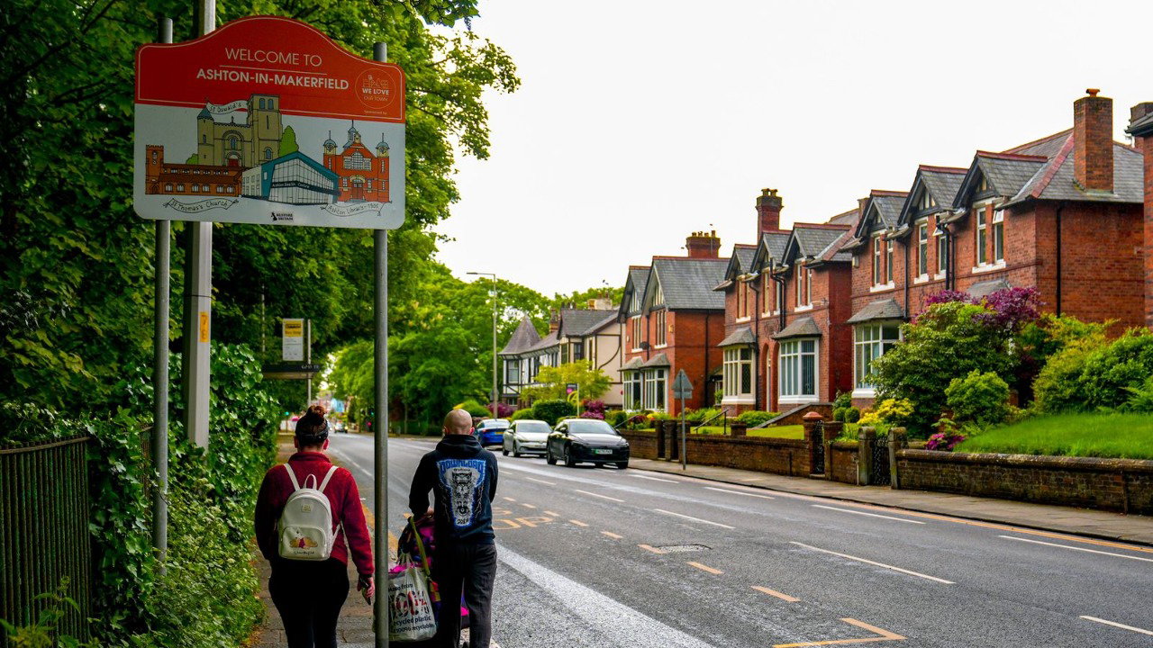 Two people with a pushchair walk on a pavement alongside a road with a bus stop. There is a sign in red with a picture of a town that says “Welcome to Ashton-in-Makerfield”. There are houses on the opposite side of the road.