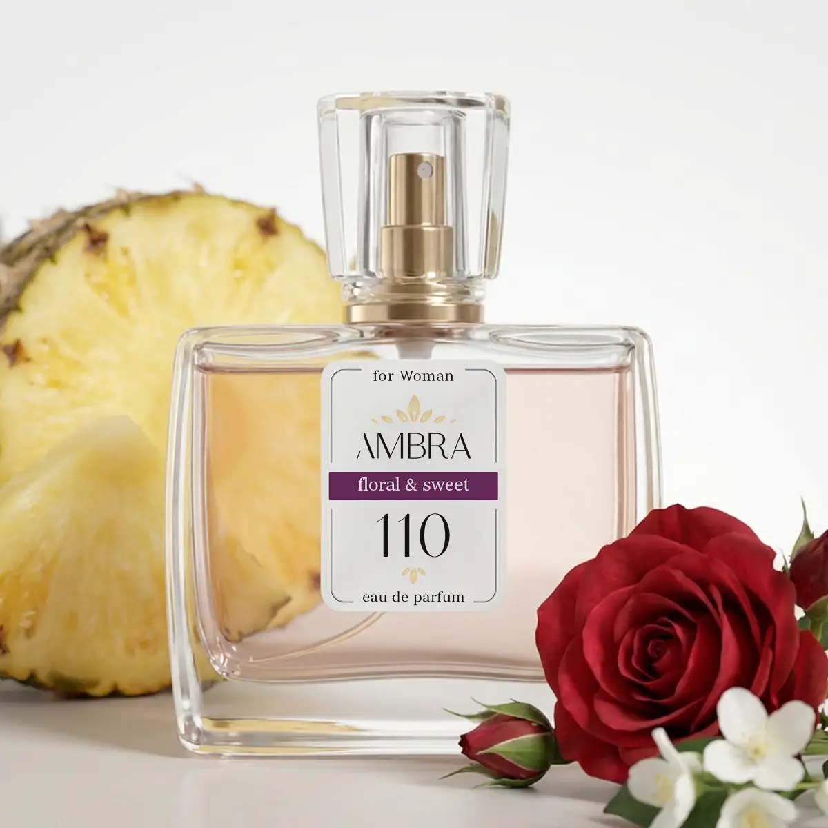 Clear glass Ambra 110 eau de parfum bottle labelled 'floral & sweet' beside a pineapple slice and a red rose