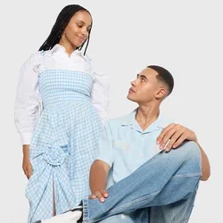 Vrouw in een blauw geruite jurk staat naast een man die op een rode stoel zit, gekleed in een lichtblauw shirt en jeans, terwijl ze naar elkaar kijken.