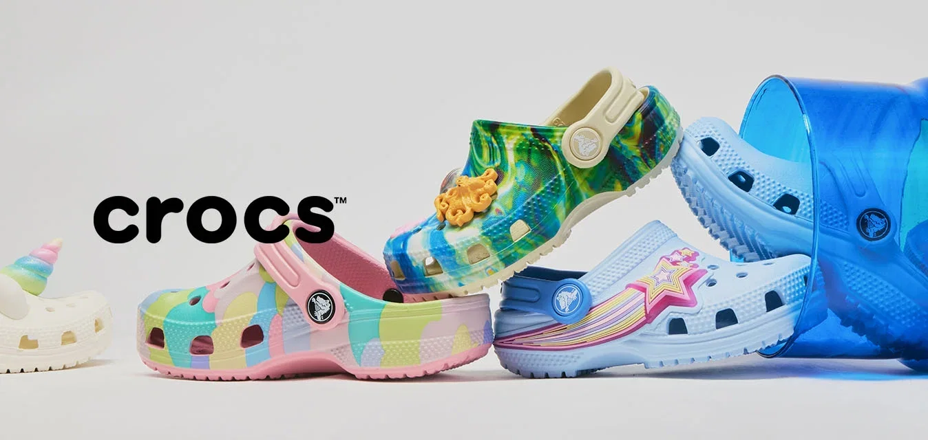 Vijf kleurrijke Crocs-schoenen speels in balans gerangschikt, waarvan twee in een omgevallen blauwe doorschijnende emmer op een witte achtergrond.