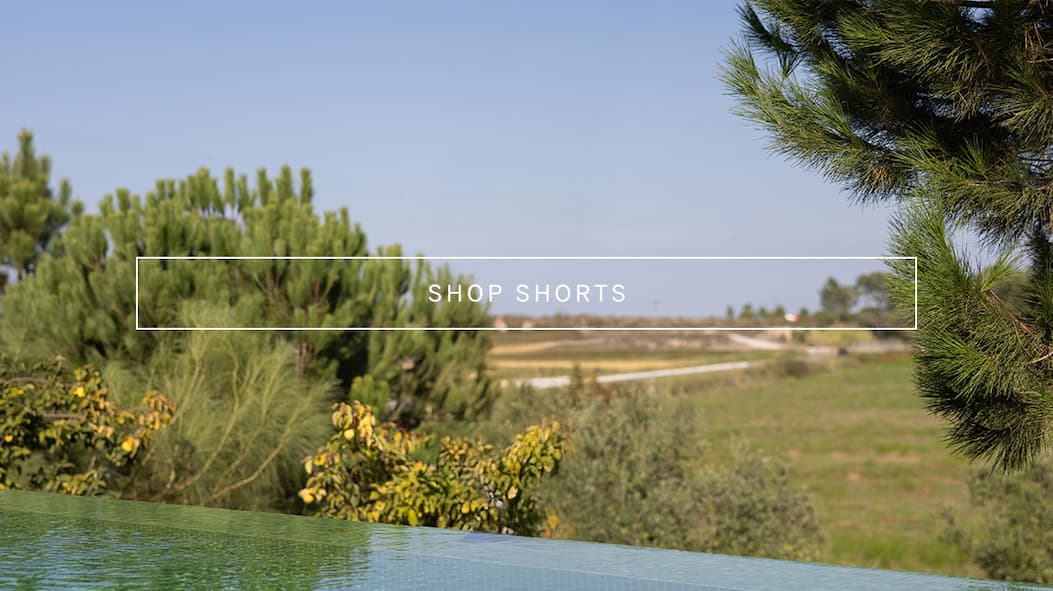 Landscape image of a poolside view surrounded by trees and greenery, with overlay text reading “Shop Shorts.”