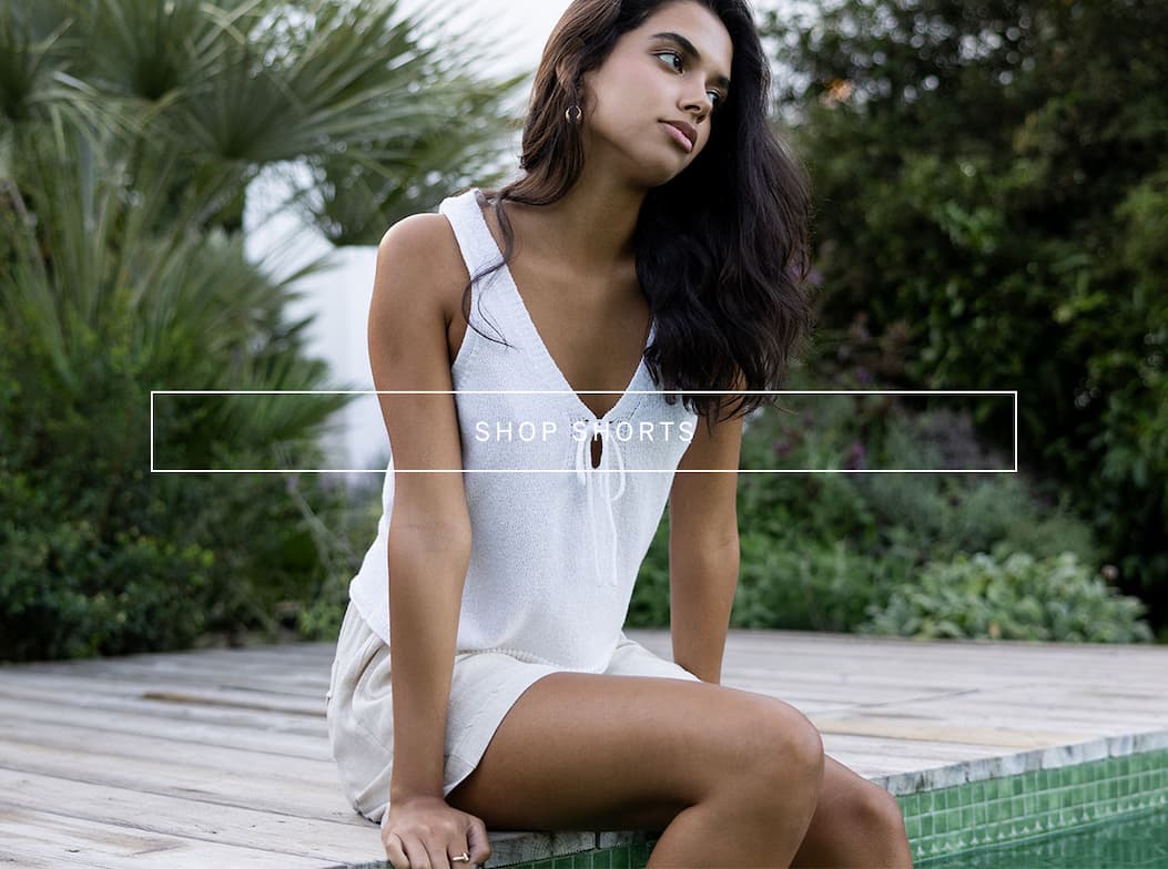 Model sitting outdoors in a white sleeveless top and light shorts beside a pool, with overlay text reading “Shop Shorts.”