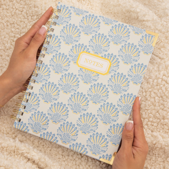 hands holding blue and gold repeated shell patterned notes book, beige blanket background