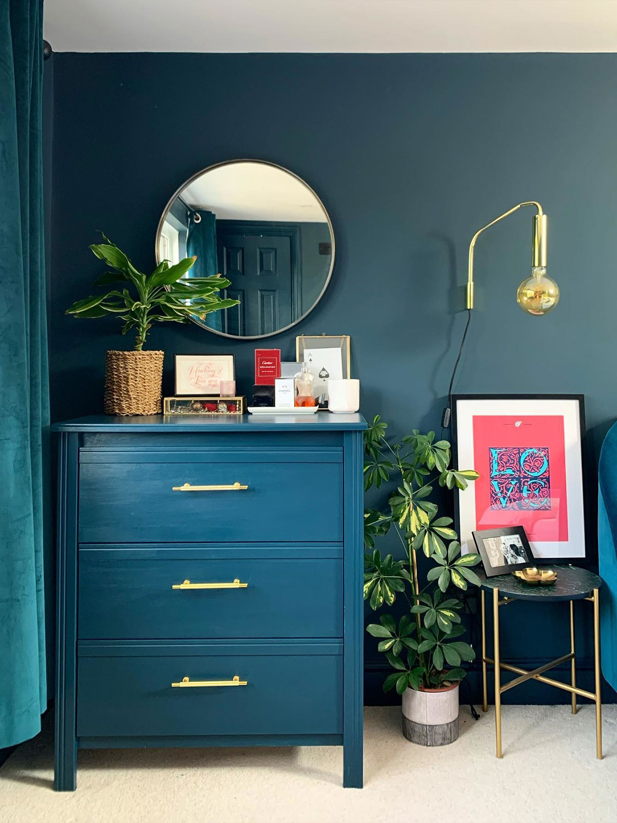 A bold teal wall and matching painted chest of drawers in a modern bedroom corner. Brass lighting, framed artwork and leafy plants bring contrast and playful energy to the space.