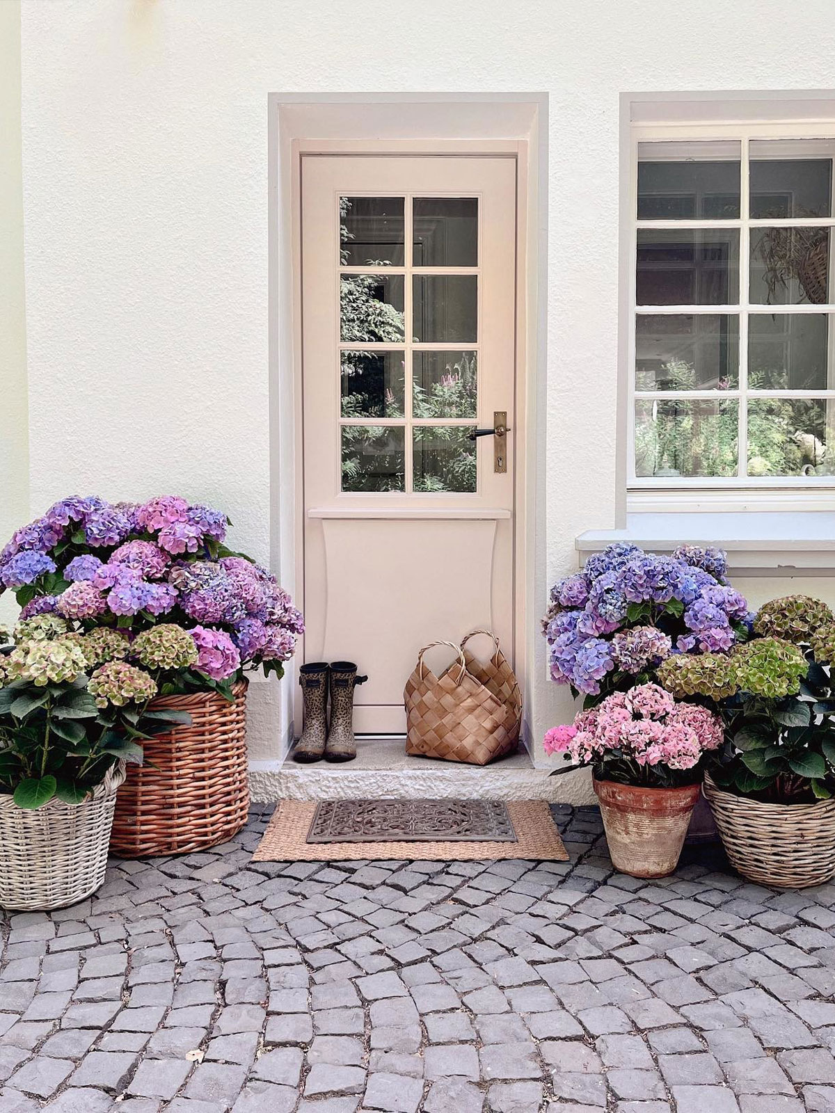 A welcoming front door framed by baskets of blooming hydrangeas, with patterned wellies and a woven doormat on a cobbled path.