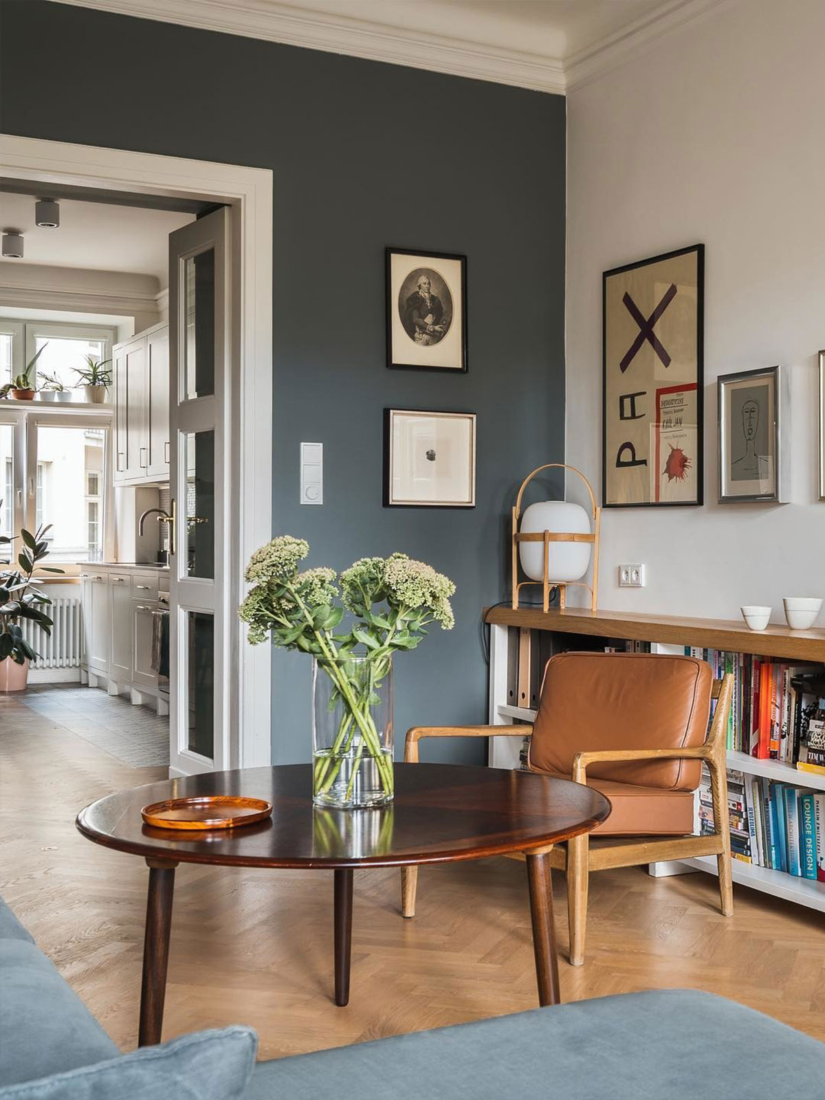 A cosy living room with a blue-grey sofa, tan armchair and a long bookshelf arranged by colour, with a view through to a bright white kitchen.