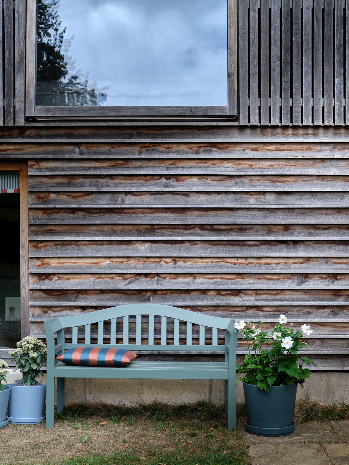 A light blue‑green bench with a striped cushion sits against a wooden exterior wall, surrounded by potted plants and a window reflecting the cloudy sky.