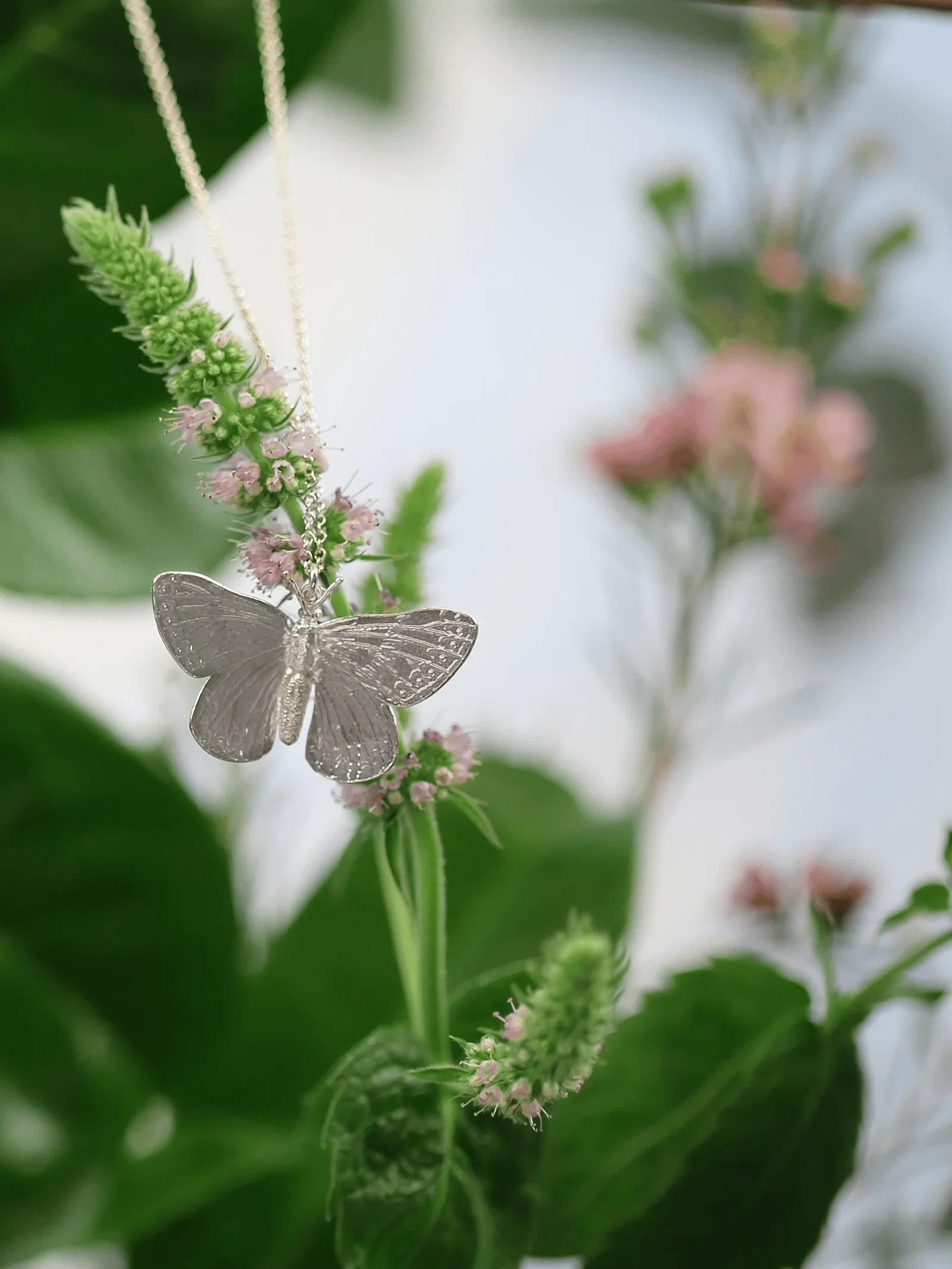 A delicate butterfly necklace hangs amongst soft wildflowers and lush green foliage in a dreamy botanical setting.