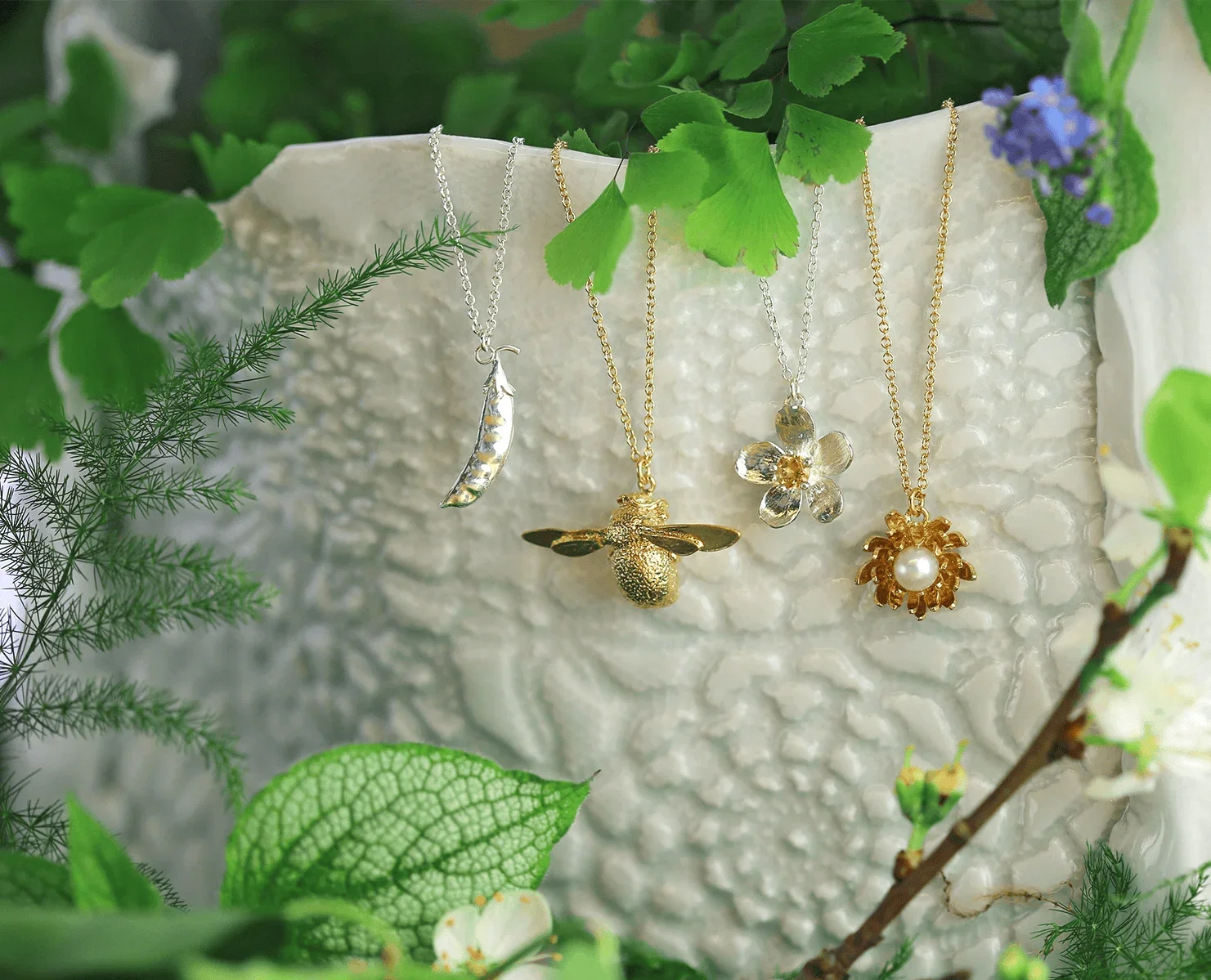 Nature-inspired necklaces drape across a textured ceramic vessel, surrounded by lush green foliage and delicate wildflowers.