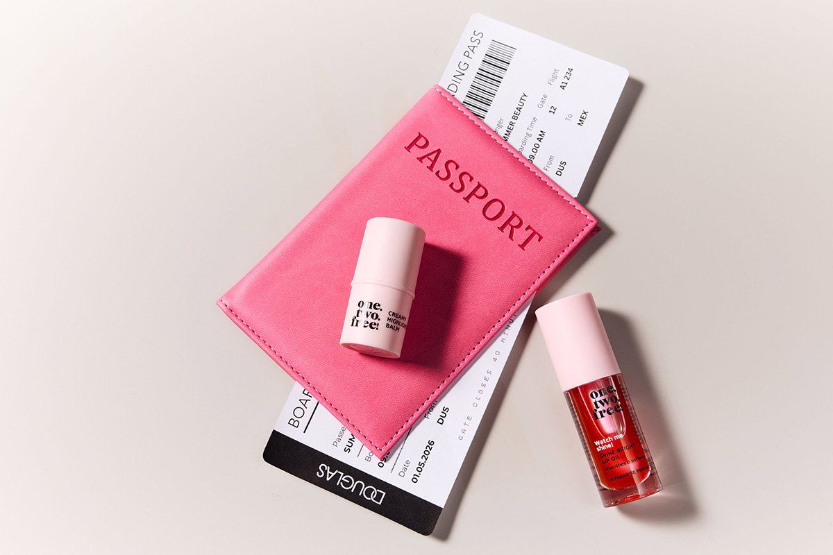 Alt-Text: „Pinke Passhülle mit Boardingpass und zwei Beauty-Produkten auf hellem Hintergrund arrangiert.“