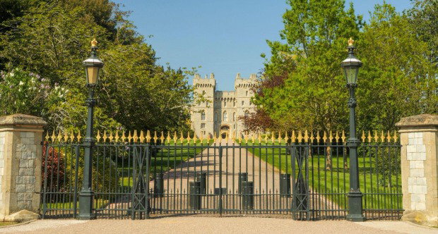 Windsor Castle Upper Ward Entrance behind gold-tipped black security gates and two lamposts. The path from the gate to the castle is bordered by a strip of green lawns and trees.