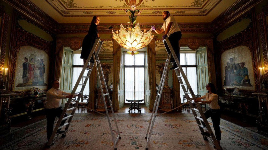 Two staff members are cleaning a massive lotus-shaped chandelier while on ladders in a grand, ornate room with high ceilings and decorative wall panels. Another two members of staff are holding the ladders at the bottom.