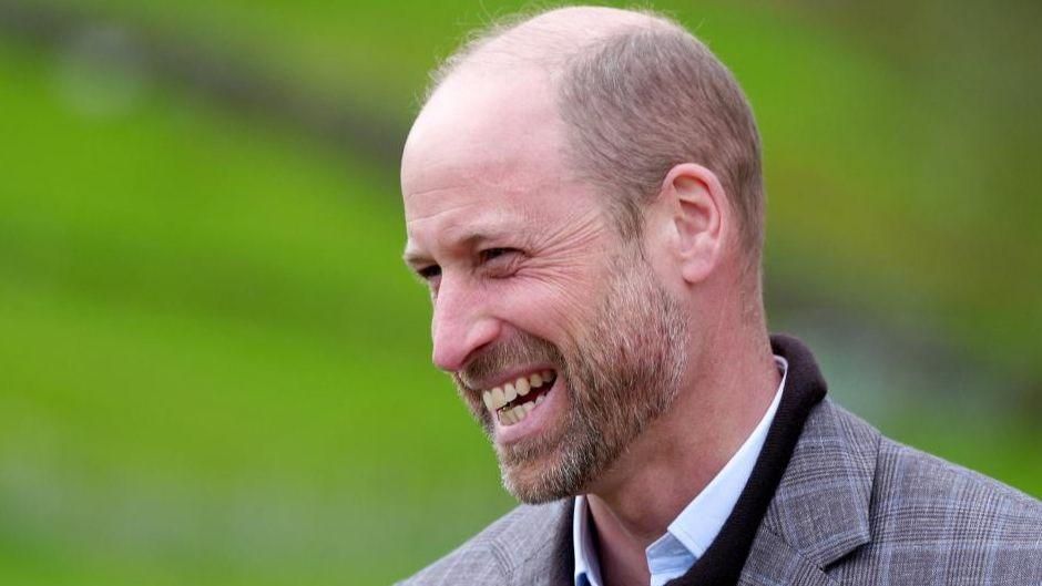 William, Prince of Wales, smiling and laughing in a grey tweed blazer against a blurred green background.