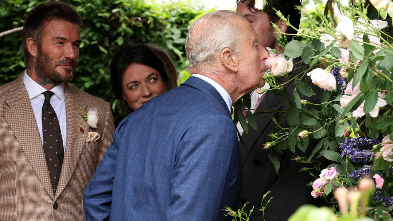 The King, wearing a blue blazer, light blue shirt and tie, is mid-laugh as he explores the garden. Next to him is the Queen wearing a black jacket with a spotted shirt collar visible at the top. She is smiling, holding a blue, purple and white bouquet of flowers. Alan Titchmarsh can be partially seen behind her, along with other visitors. David Beckham, wearing a beige suit, light shirt and brow tie, is next to the Queen, laughing and looking at the flowers.