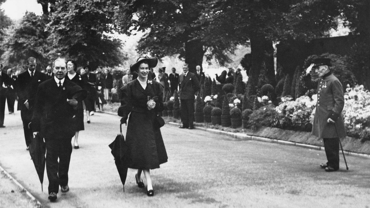 A black and white photo of a young Queen Elizabeth II smiling and walking along a garden path with others, holding a closed umbrella and wearing a dark coat and hat, while people follow behind and a man in uniform salutes nearby.