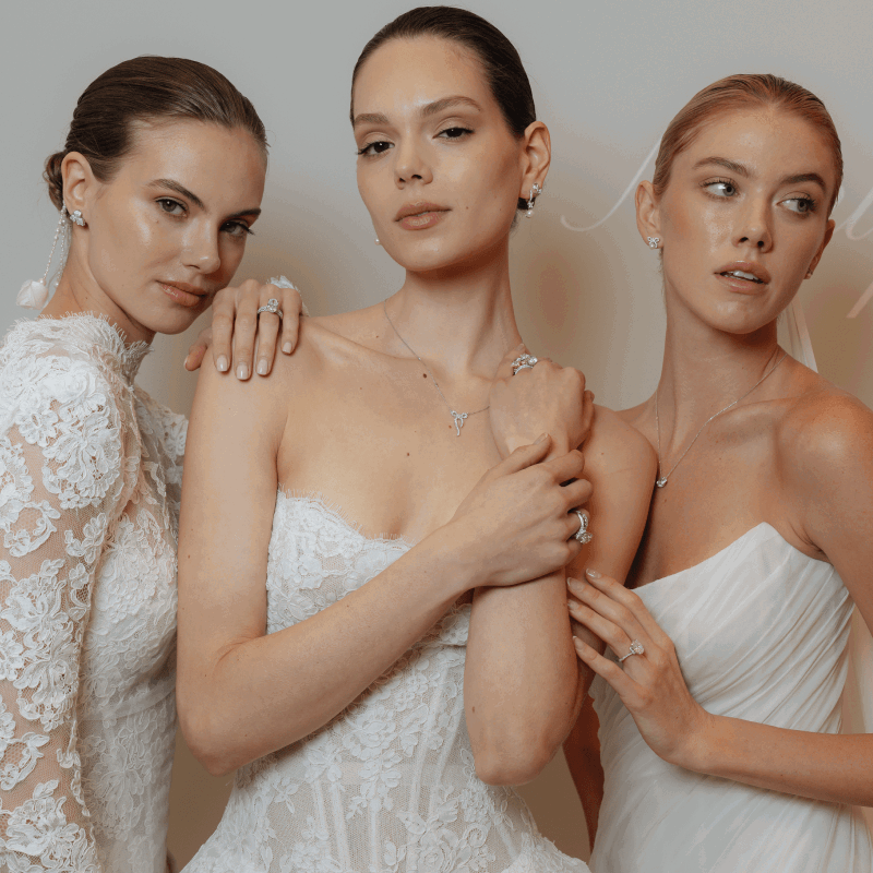 Three women in white dresses wearing diamond rings, earrings, and necklaces posing together.