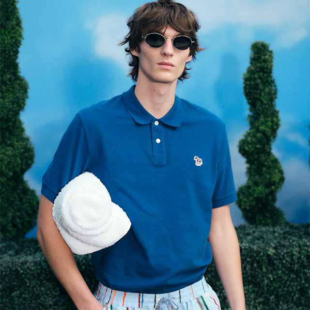 A person wearing sunglasses and a blue polo shirt holds a white bucket hat. The background features trimmed bushes and a blue sky.
