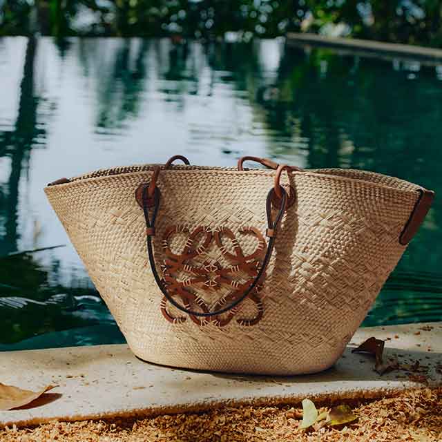 A woven straw bag with leather handles is placed on a patio near a pool and foliage.