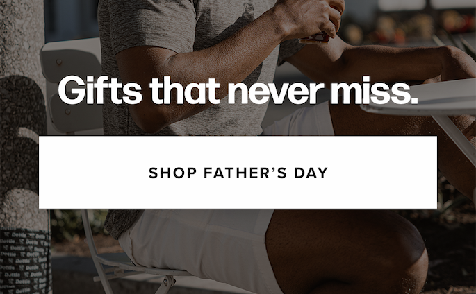 A cropped shot of a man wearing a grey t-shirt and white shorts while sitting at an outdoor table and holding a beverage. Text reads: 'Gifts that never miss. Shop Father’s Day'