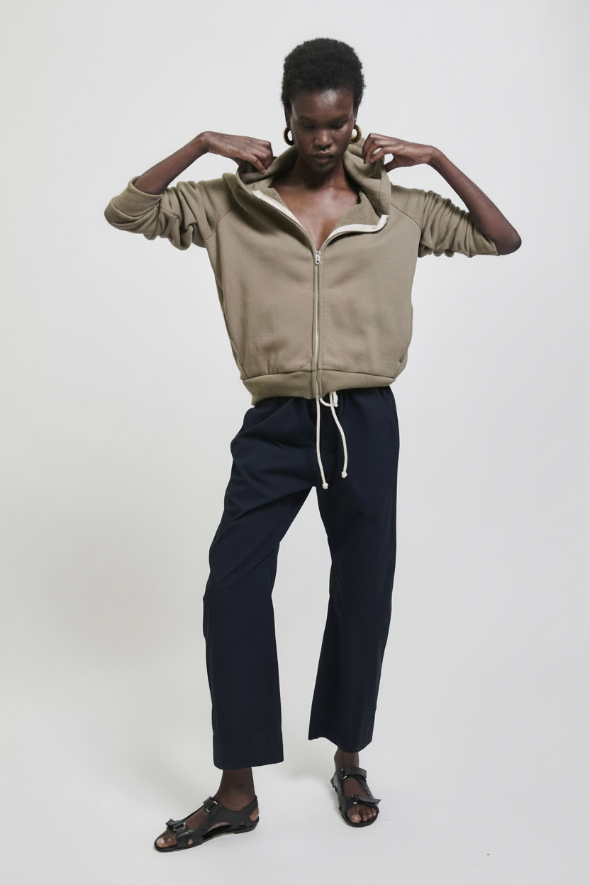A woman in a tan zip-up hoodie and navy pants adjusts her hood.