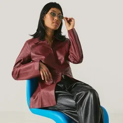 Frau mit einer kastanienbraunen Jacke und schwarzen Hosen sitzt auf einem modernen blauen Stuhl und richtet ihre Sonnenbrille vor einem schlichten hellen Hintergrund.