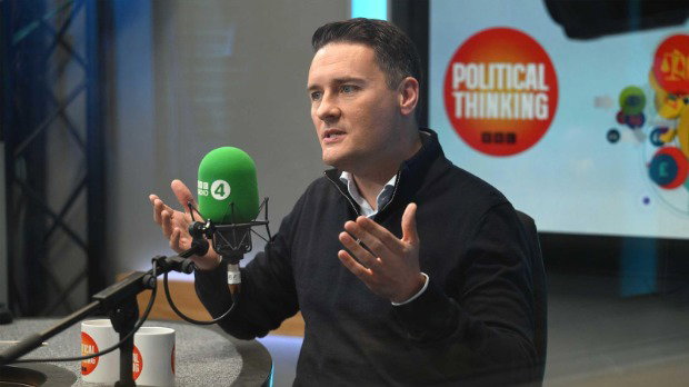 Former Health Secretary Wes Streeting giving an interview to the BBC's Political Thinking podcast, wearing a dark sweater. He raises his hands as he speaks into a green microphone, which says ‘BBC Radio 4’ in white lettering. Blurred in the background is a ‘Political Thinking’ logo.